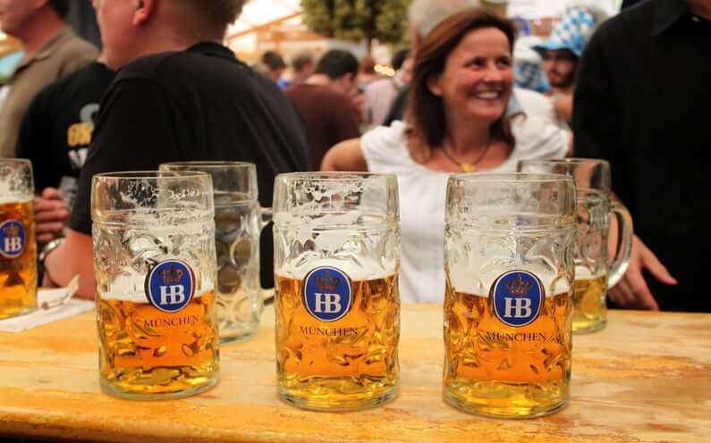 Three half-full mugs of beer with blue labels sit on a wooden table in a crowded beer garden.