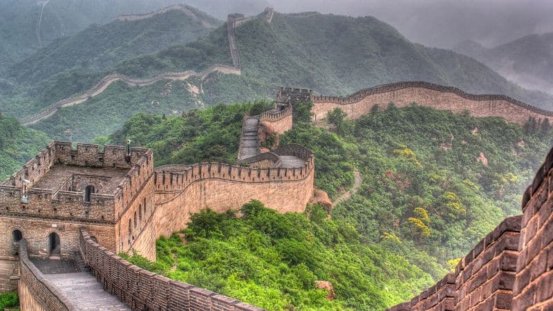 An aerial shot shows the Great Wall of China snaking through green forests. The wall is made of brick and has two square turrets in frame.