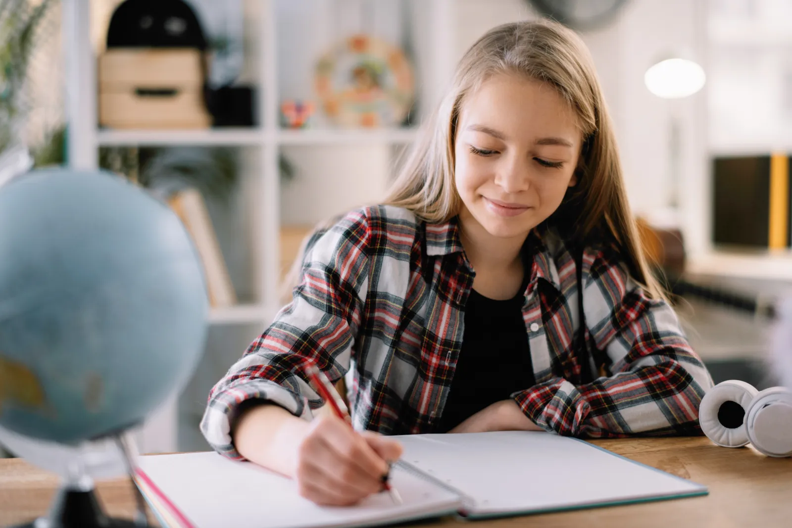 A student writing in a notebook at a desk, a globe and headphones beside her.