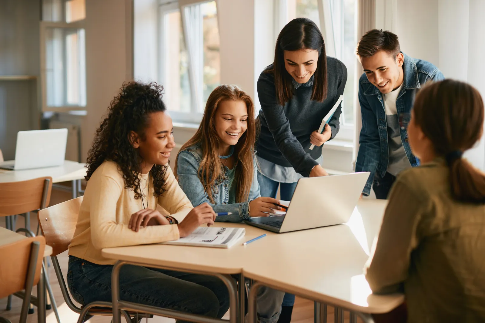 A teacher leaning over a table of teenagers working at laptops, talking them through something on screen.