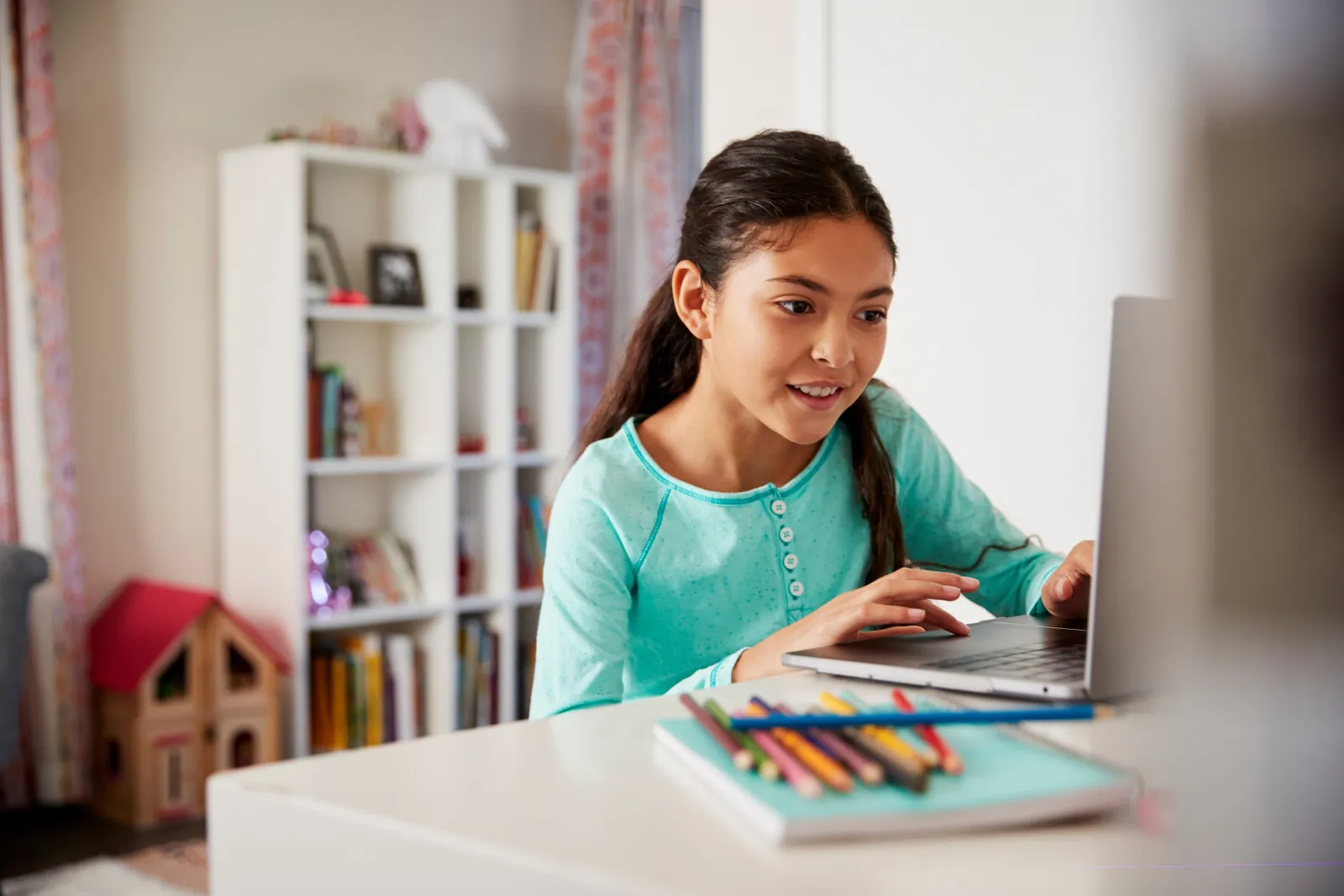 A girl smiling at a laptop at her bedroom desk, coloured pencils and a bookshelf beside her.