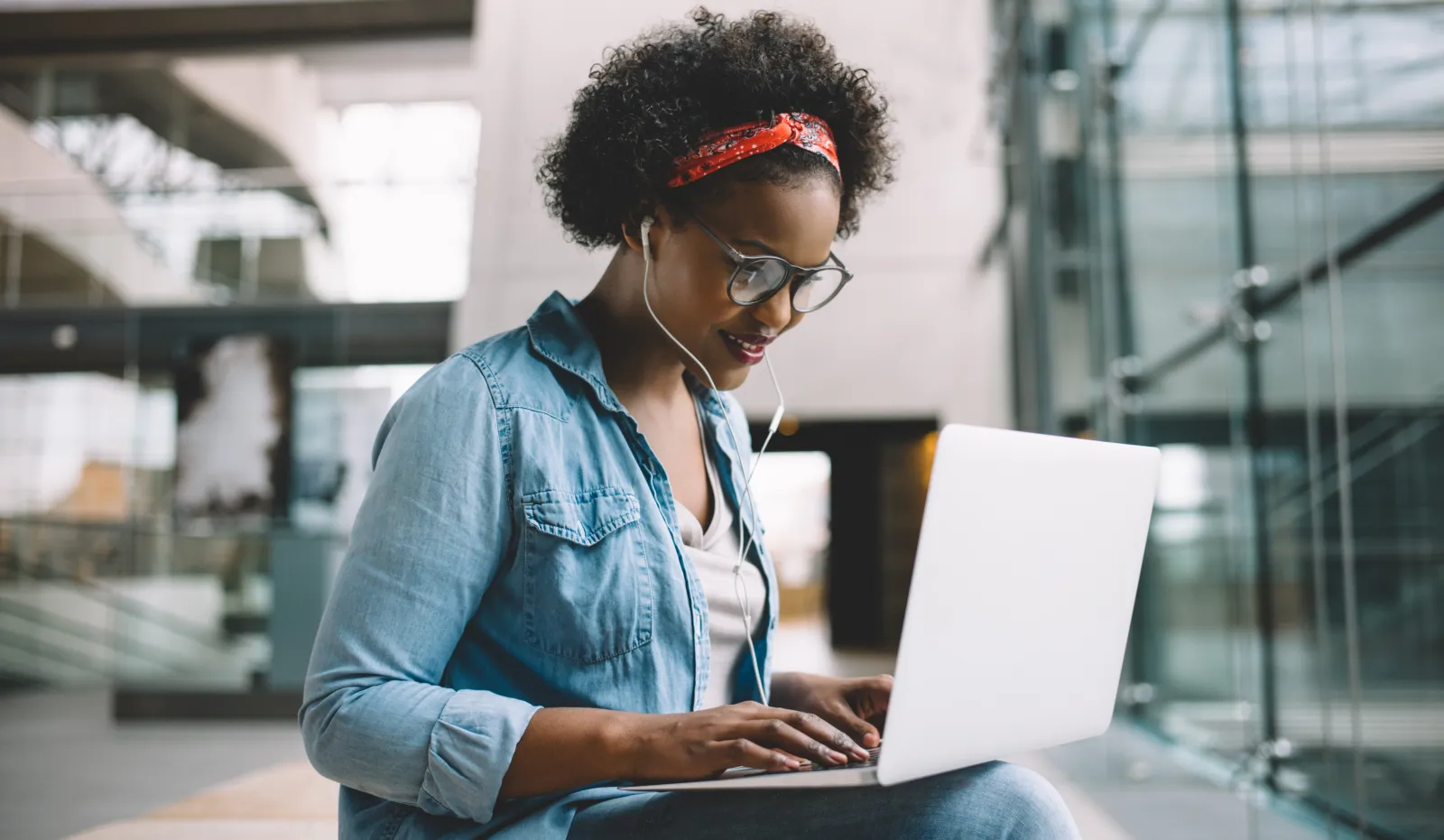 A woman with earphones in working at a laptop on a step outside a glass building.