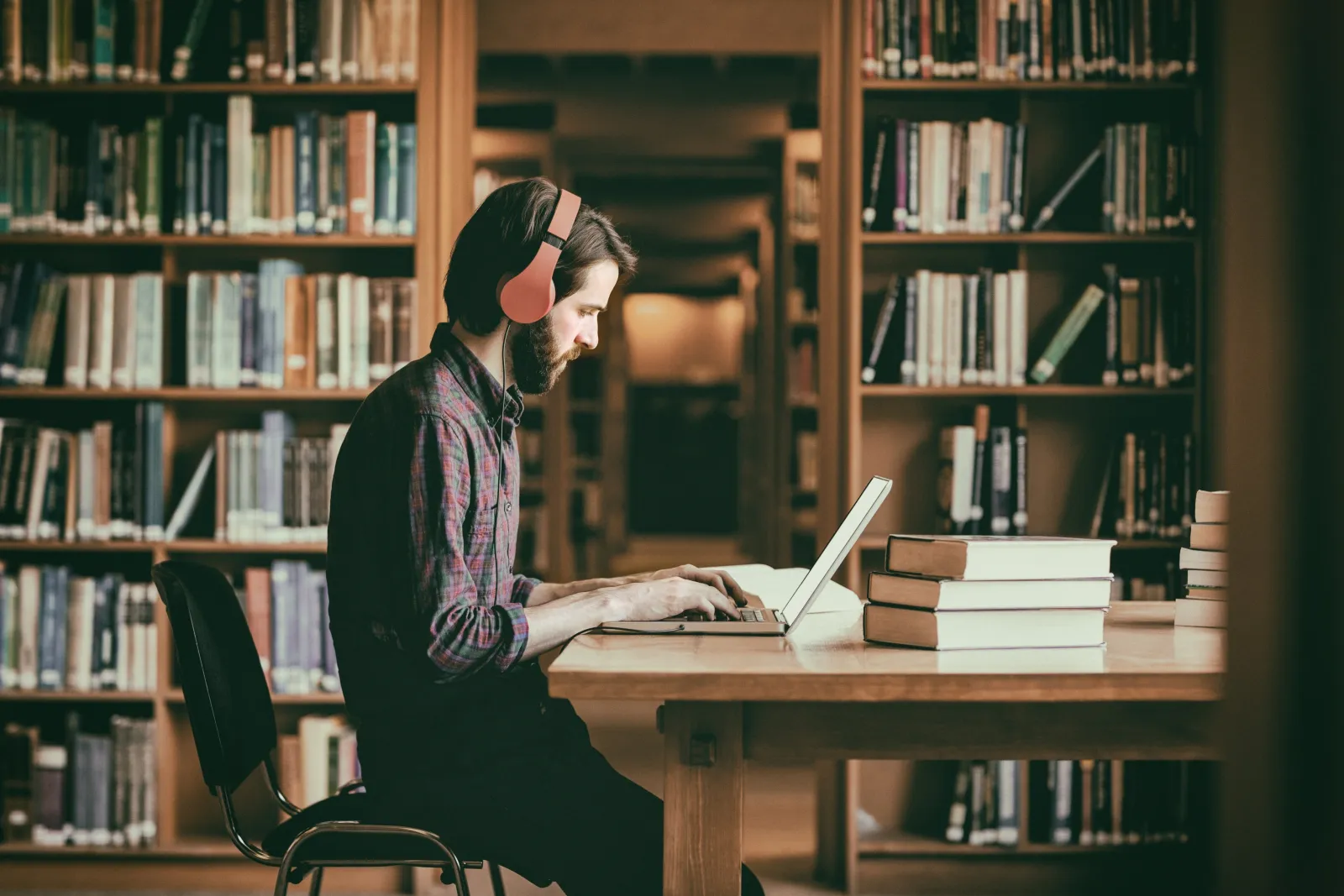 A man in headphones reading at a desk between tall dark wooden bookcases.