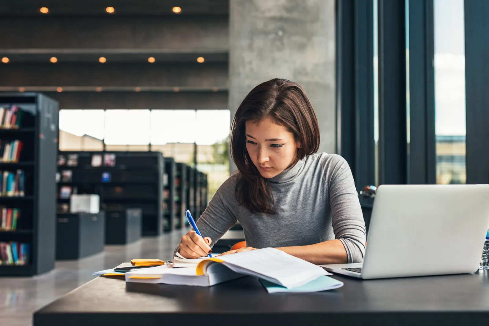 A student writing in a notebook beside an open laptop at a library desk.