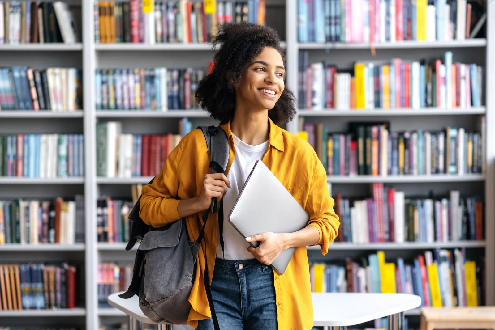 A smiling student in a yellow jacket carrying a laptop past tall library bookshelves.