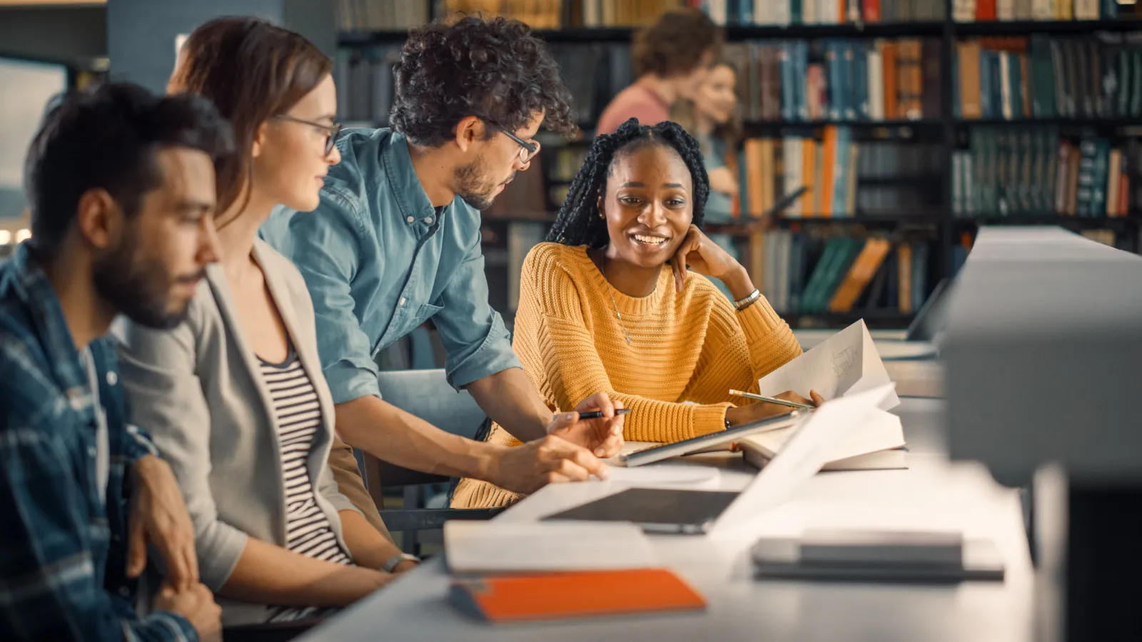 Students gathered around a library table with laptops and notes, talking over their work.