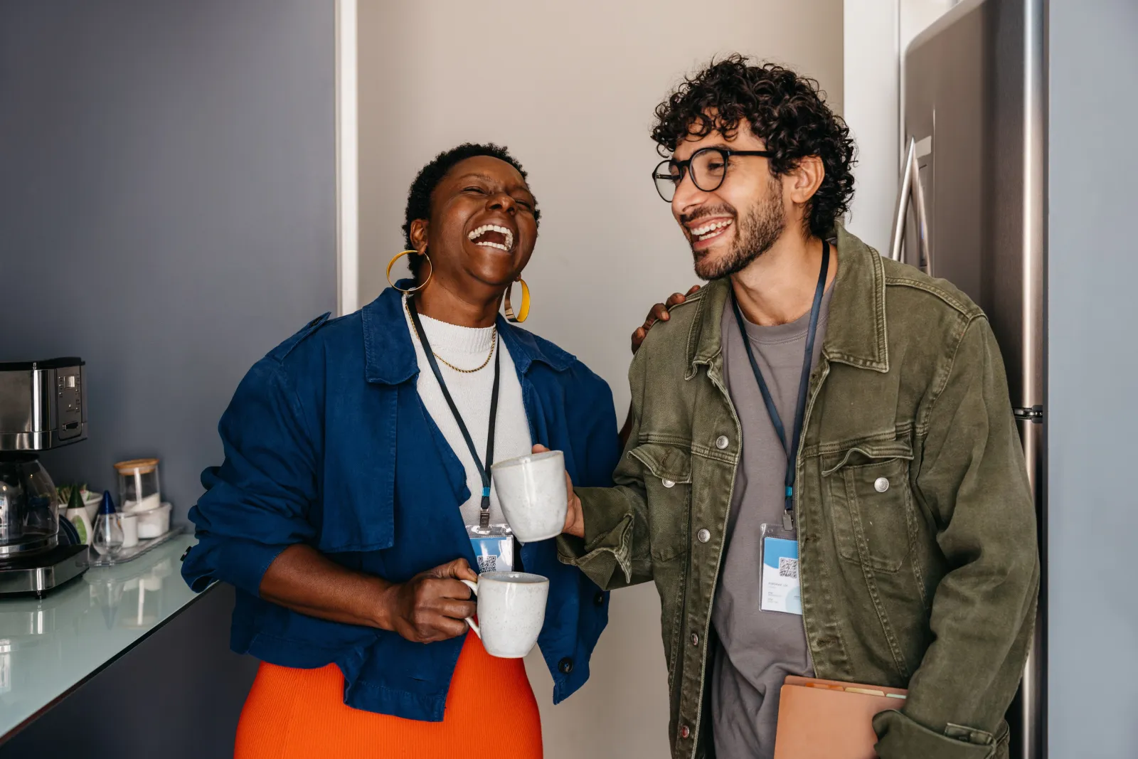 Two colleagues laughing together over coffee, lanyards around their necks.