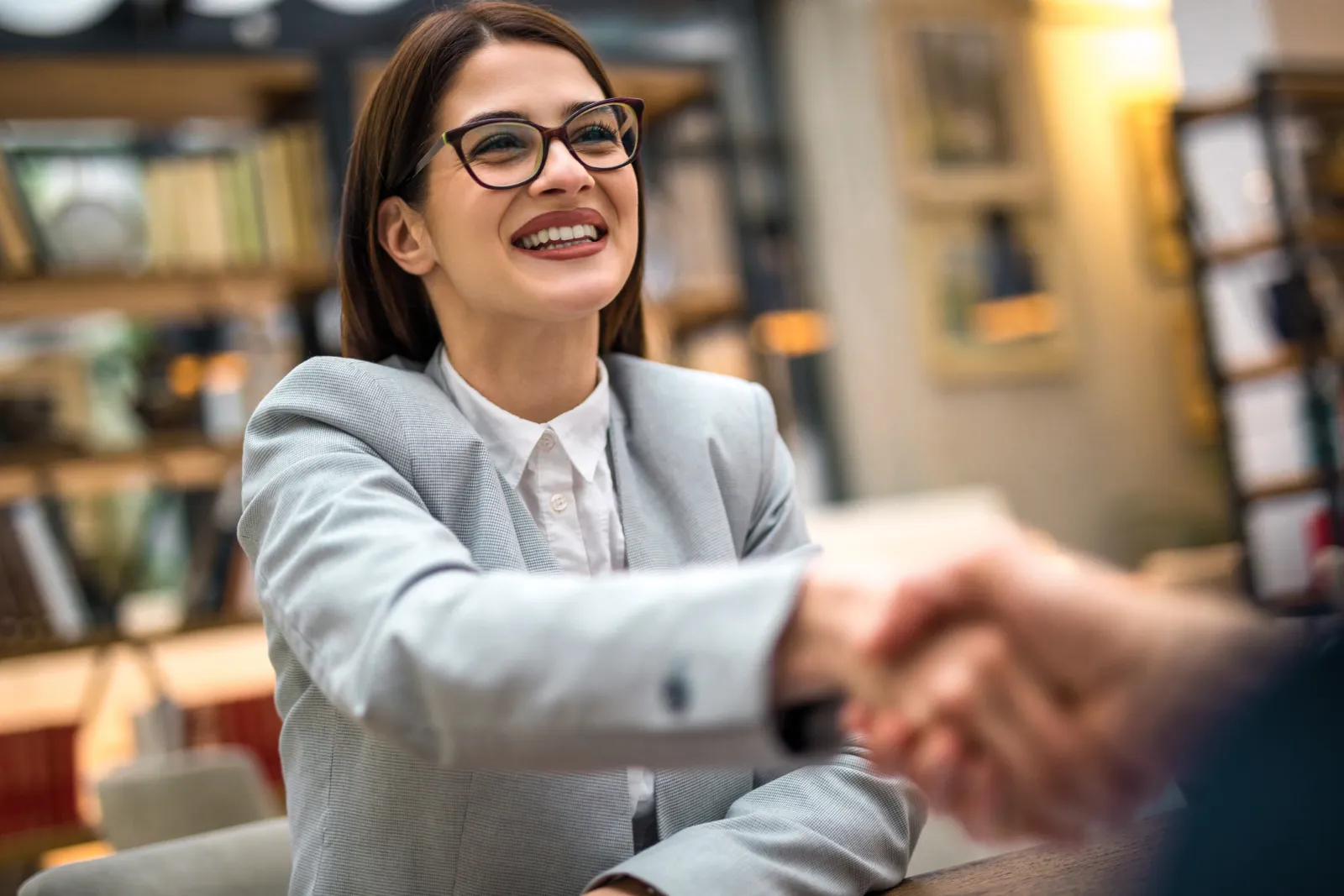 A woman in glasses smiling as she shakes hands with someone across a desk.