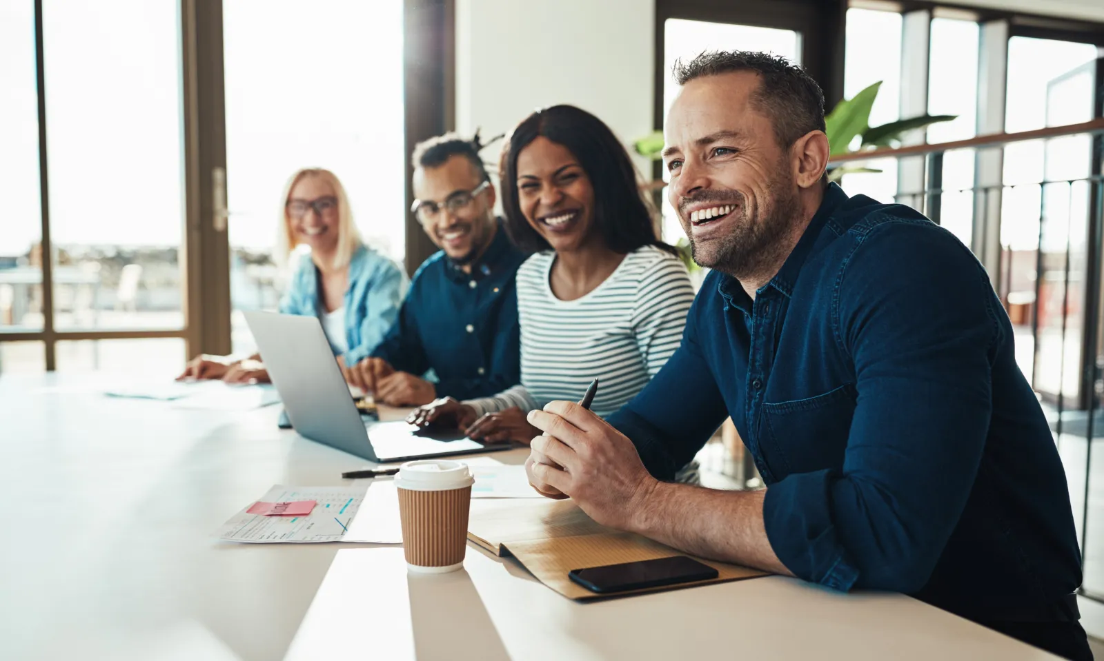 Four colleagues smiling around a meeting table with open laptops, listening to someone out of frame.